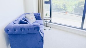 A room with a blue sofa, a small round table, and a view of a road and greenery through a large window. - Heart of AKL Modern Studio PoolGymSauna (Auckland)