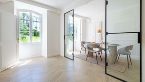A spacious room with a herringbone wooden floor, a dining area with a round table and chairs, and a large sliding glass door leading to a garden. - Garden-Level Apartment in Bourgeois Residence – Near Croisette & Beaches (Cannes)