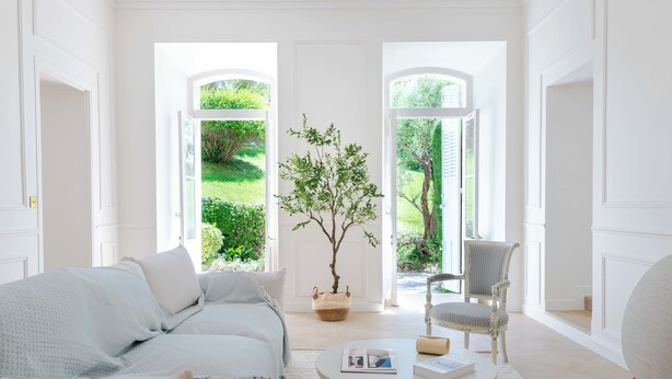 A bright, white room with a sofa, a round coffee table, and a chair. There are two large windows with a view of greenery outside. - Garden-Level Apartment in Bourgeois Residence – Near Croisette & Beaches (Cannes)