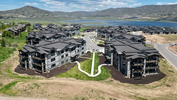 A residential area with modern multi-story buildings, a winding road, and a lake in the background. - Mayflower Lakeside 103 | Mountain View | Balcony | Full Kitchen | Private Bedrooms | Fireplace (Heber City)