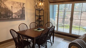 A dining area with a wooden table and chairs, a chandelier, a bookshelf, and a large window with a view of trees.