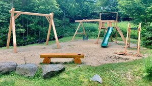 A playground with swings, a slide, and a bench in a wooded area.