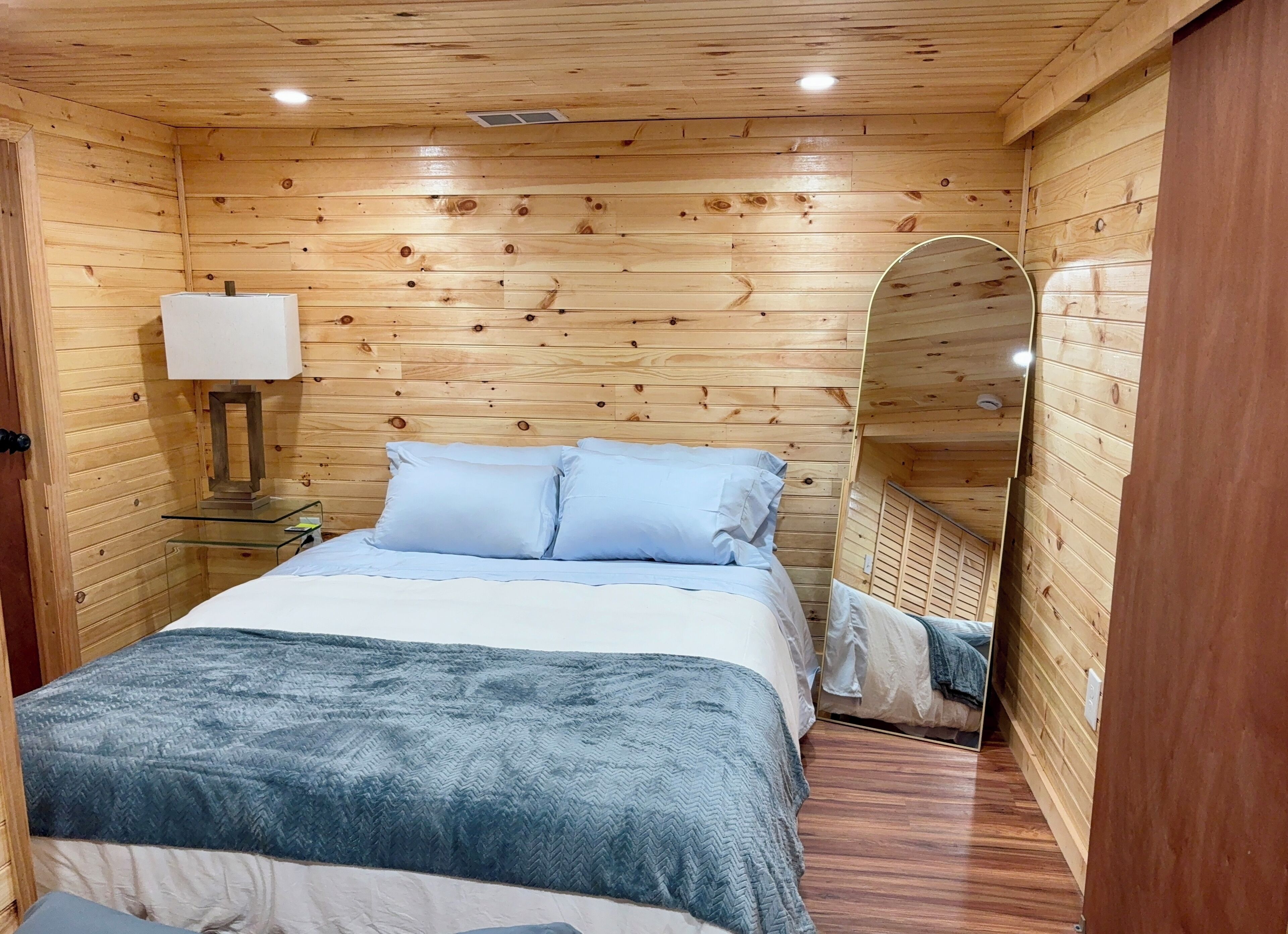 A wooden-paneled room with a bed, a bedside table with a lamp, and a mirror.