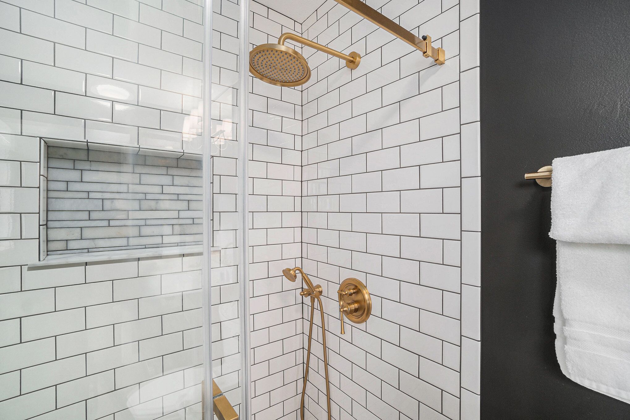 Une douche dotée d’un pommeau de douche doré et d’un pommeau de douche à main, des murs carrelés de blanc et de noir, et une serviette blanche accrochée au mur.