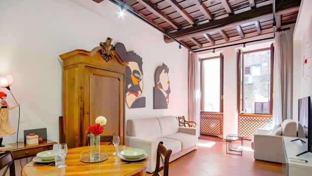 A dining area with a wooden table, chairs, a cabinet, and a vase with flowers. There is a white sofa and a balcony with a railing. - Elegantis.it Eat Pray and Love in Rome - Near Navona Square and Campo de' Fiori (Roma)