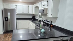 A modern kitchen with white cabinets, a stainless steel refrigerator, a black countertop, and a bar area with stools. -  Welcome to mountain retreat in the tranquil beauty of Rabun County! (Clayton)