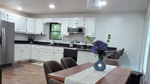 A modern kitchen with white cabinets, a wooden dining table with chairs, and a hanging light fixture. -  Welcome to mountain retreat in the tranquil beauty of Rabun County! (Clayton)