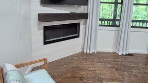 A living room with a flat-screen TV mounted on the wall, a wooden chair with cushions, a patterned rug, and a window with curtains. -  Welcome to mountain retreat in the tranquil beauty of Rabun County! (Clayton)