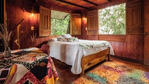 A wooden room with a bed, a table with a patterned tablecloth, and a window with a view of trees. - Tominejo Ecolodge (Neira)