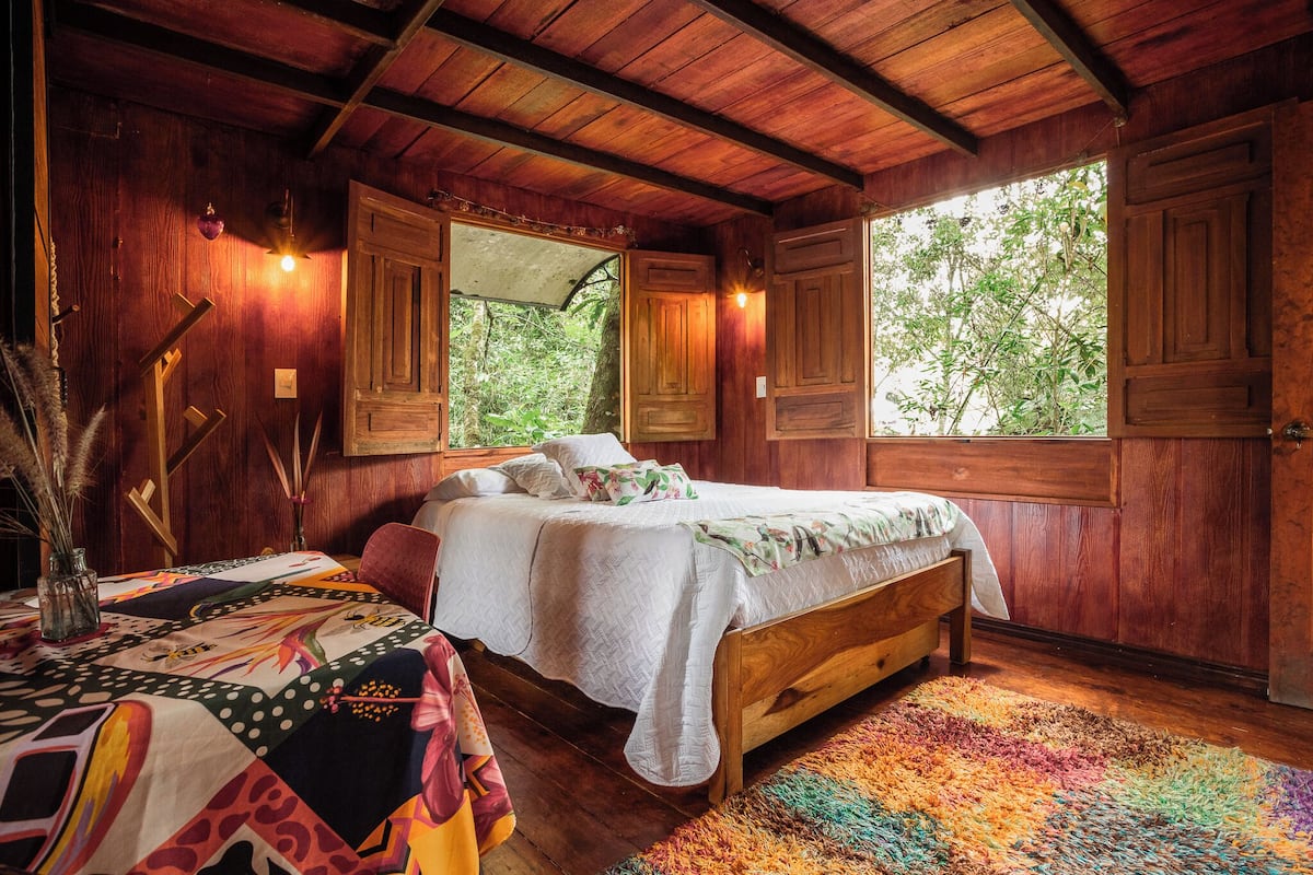 A wooden room with a bed, a table with a patterned tablecloth, and a window with a view of trees.
