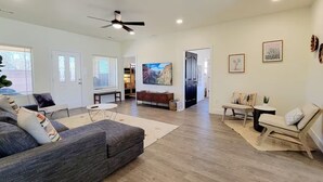 A spacious living room with a grey sofa, a small round table, and a bench. There is a ceiling fan, a large painting on the wall, and a door leading to another room. - Your Kanab Retreat w/ Hot Tub, Game Room, Pet Friendly (Kanab)