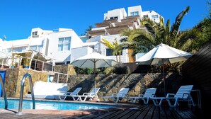 Zona de piscina con sillones de descanso y sombrillas, un muro de piedra y un edificio con varios balcones.