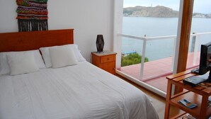 Un dormitorio con una cama grande, una mesita de noche de madera, un tapiz colgado en la pared y vista al mar desde un balcón.