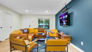 A modern living room with a sectional sofa, yellow armchairs, a coffee table, and a flat-screen TV mounted on a blue wall. - LUX Backyard Oasis, Pool, OutdoorTV, Covered Patio, Loungers, Walk to Stadiums (Arlington)