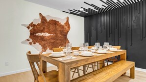 A dining area with a wooden table set for a meal, a large wall-mounted animal hide, and a modern black wall with vertical lines. - LUX Backyard Oasis, Pool, OutdoorTV, Covered Patio, Loungers, Walk to Stadiums (Arlington)