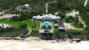 An aerial view of a large, white, multi-story building with a swimming pool, surrounded by lush greenery and a beach.