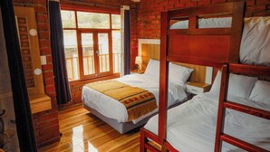 Una habitación con pared de ladrillo, una cama litera de madera, una cama individual con colcha blanca, una ventana con cortinas y suelo de madera.