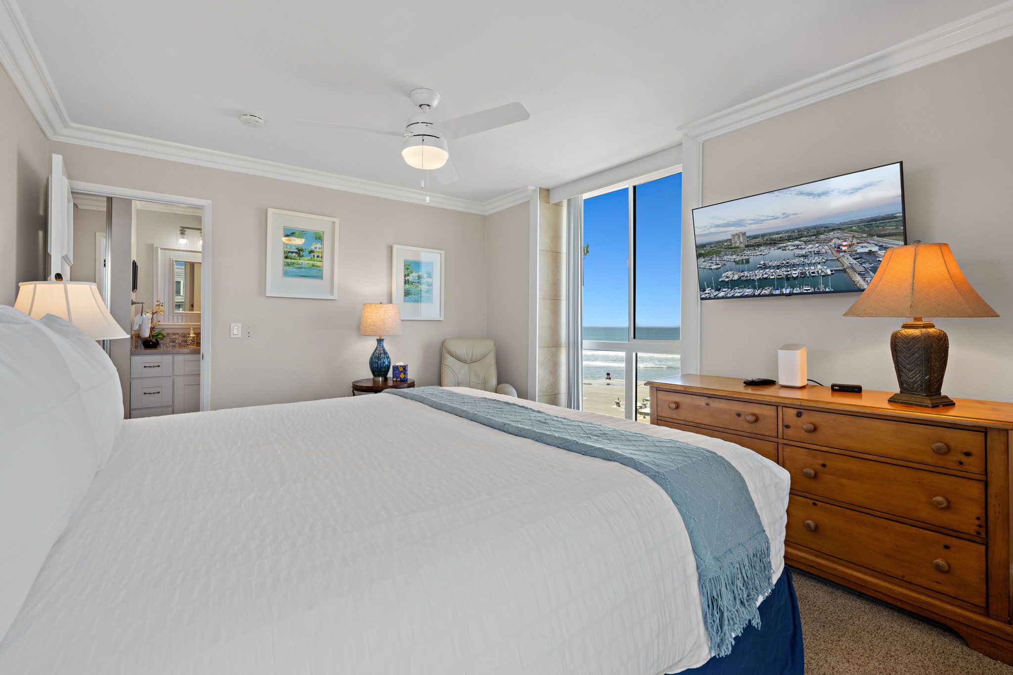 A hotel room with a large bed, a desk with a lamp, a chair, a dresser, and a television displaying a marina view.