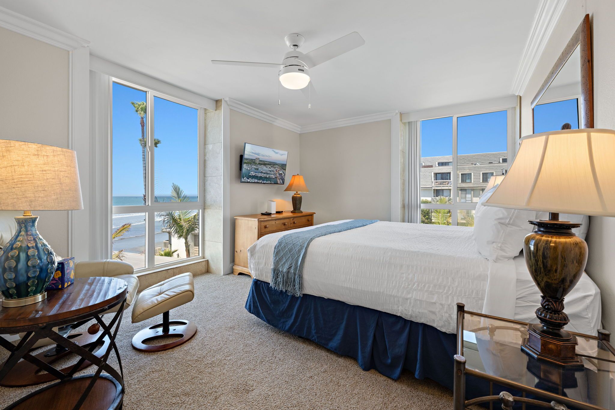 A hotel room with a bed, a desk, a chair, a TV, and a view of the beach.