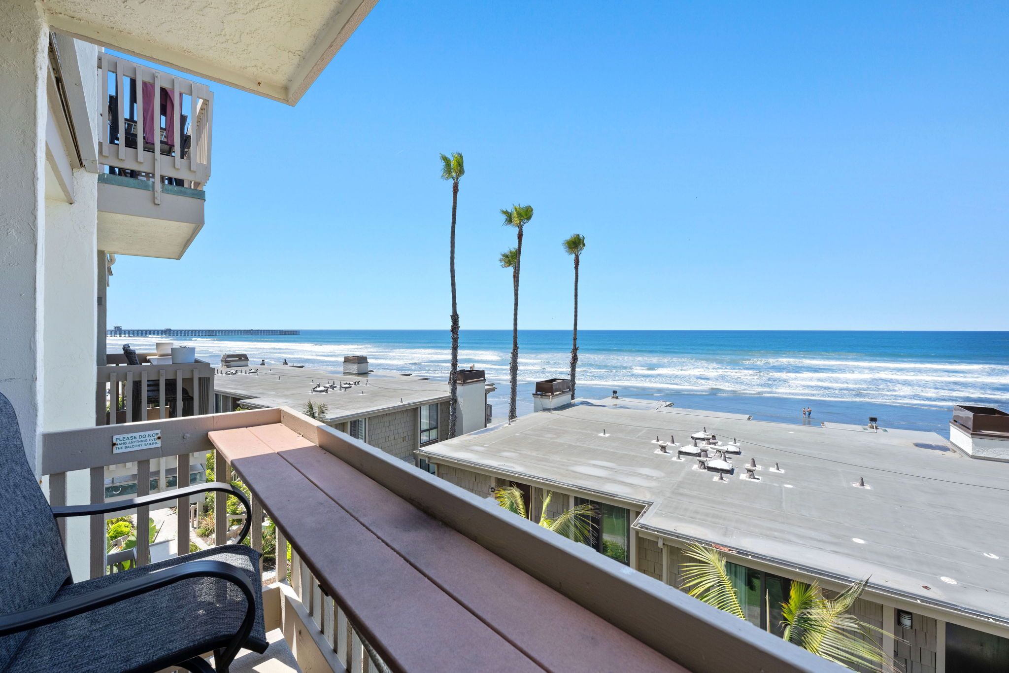 Ein Balkon mit Blick auf Strand und Meer, ein Parkplatz sowie ein Schild, das vor hängenden Gegenständen am Balkongeländer warnt.