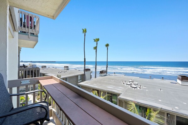 A balcony with a view of the beach and ocean, a parking area, and a sign warning against hanging objects over the balcony railing.