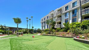 Une cour bien entretenue, dotée d’un green d’entraînement au golf, entourée d’immeubles à plusieurs étages avec des balcons, le tout sous un ciel bleu azur.