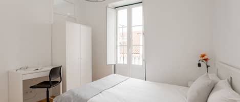 A bedroom with a bed, a desk, and a chair. There is a window with a view of buildings outside.