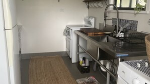 A compact kitchen with a refrigerator, stove, sink, and washing machine. There are shelves with kitchenware and a rug on the floor.