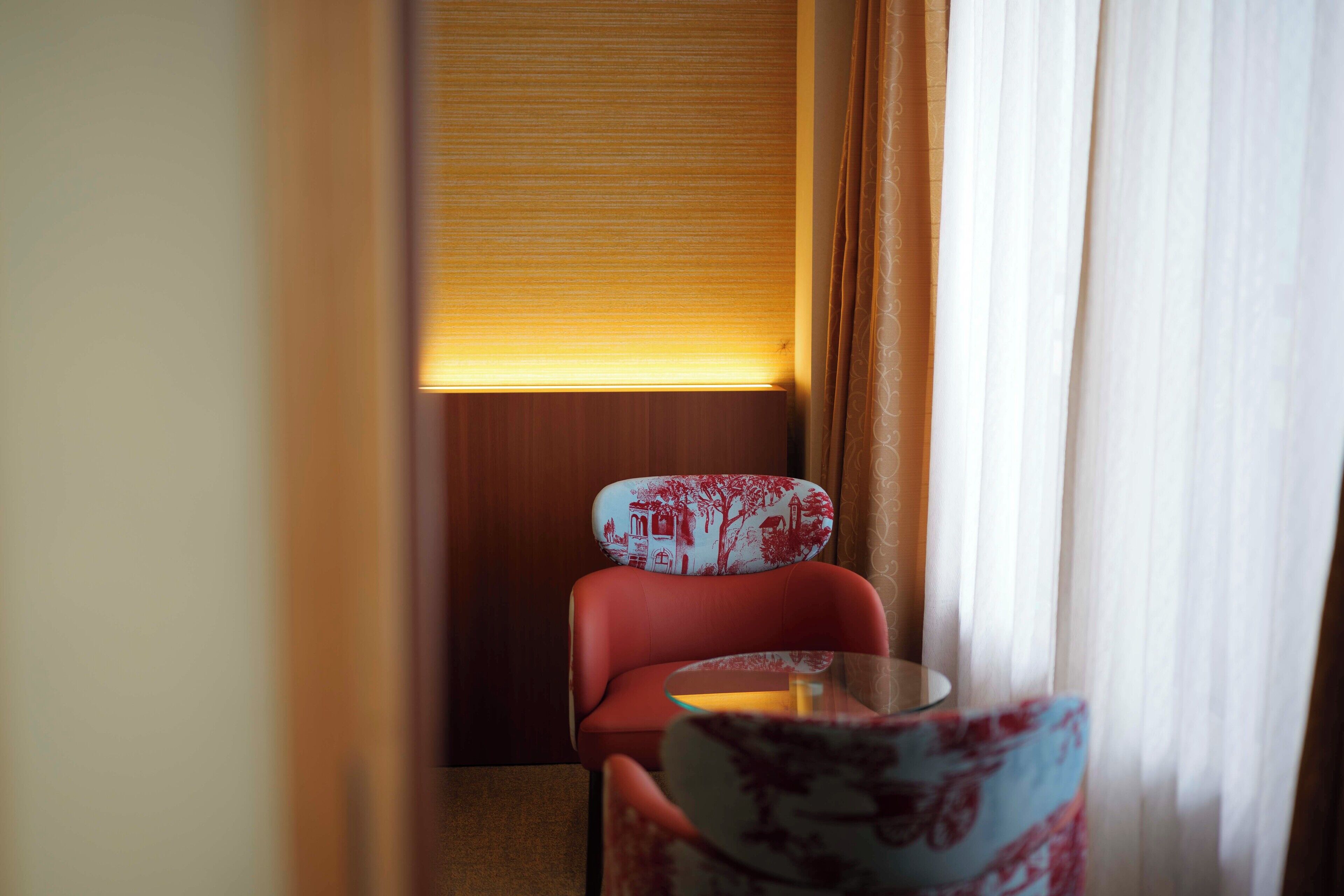 A hotel room with a red chair, a small table, and a window with curtains.