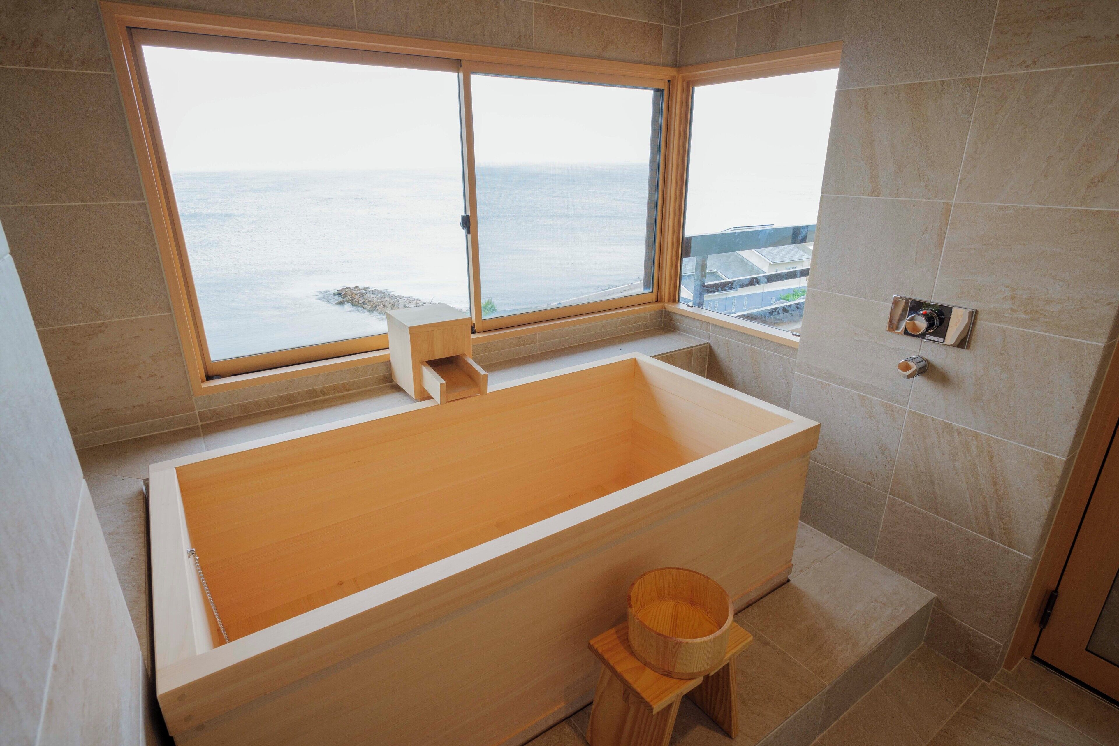A modern bathroom with a large bathtub, wooden accents, and a view of the sea through large windows.