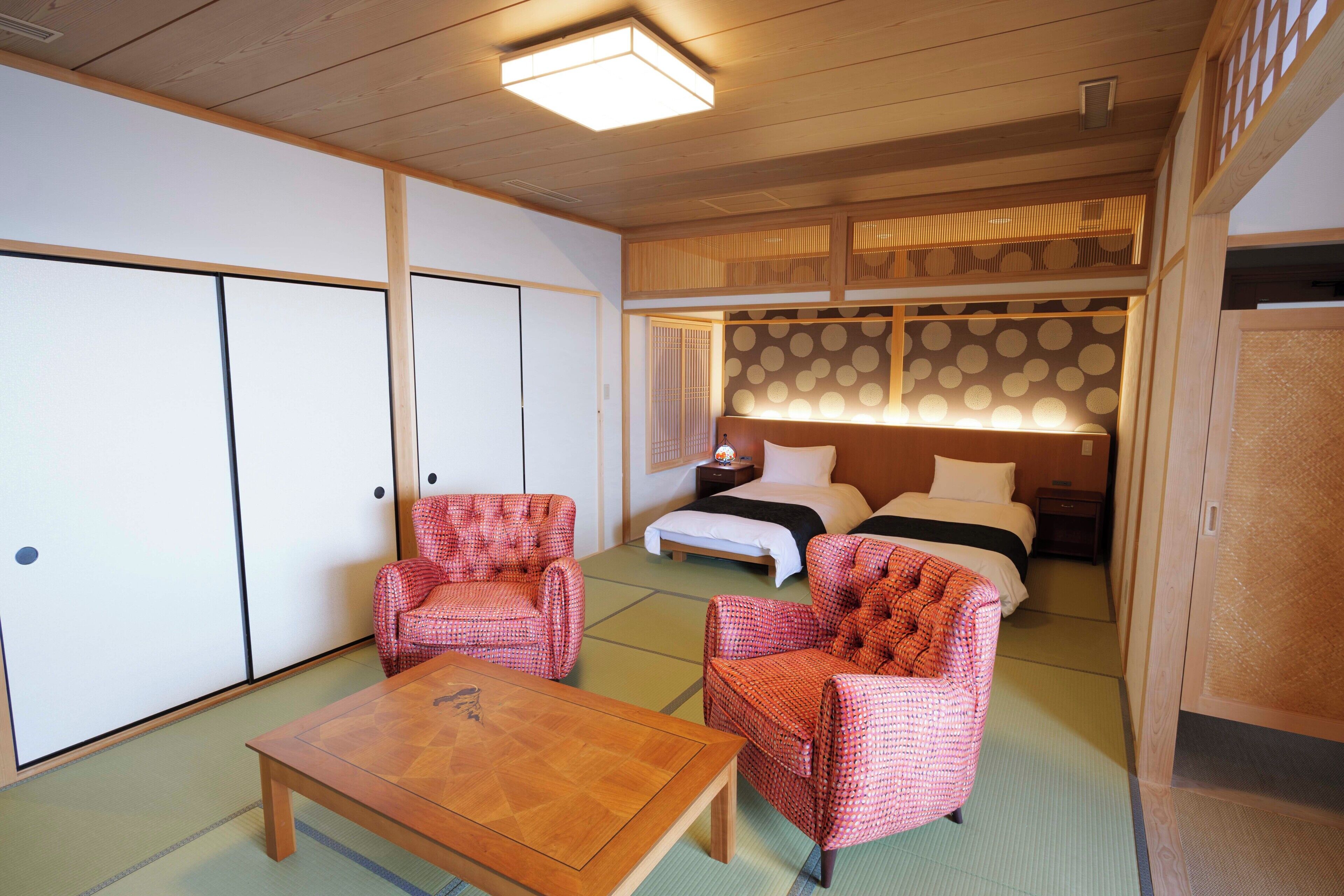A traditional Japanese-style room with tatami flooring, a low wooden table, two red patterned chairs, a bed with a white and black bedspread, and a sliding shoji screen with circular patterns.