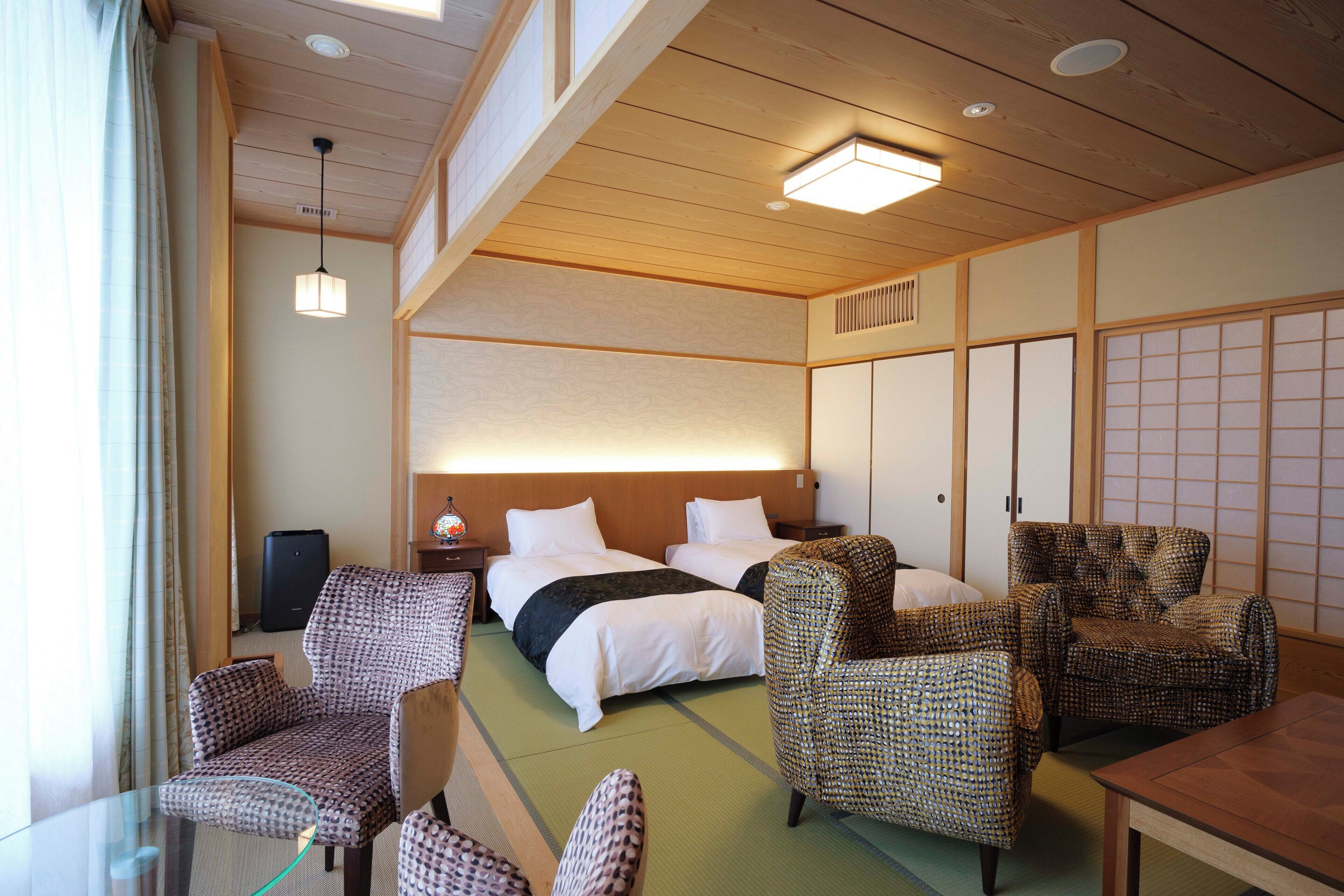 A traditional Japanese-style hotel room with tatami flooring, a low wooden ceiling, and shoji screens. There are two beds with white bedding, a small table, and several patterned chairs.