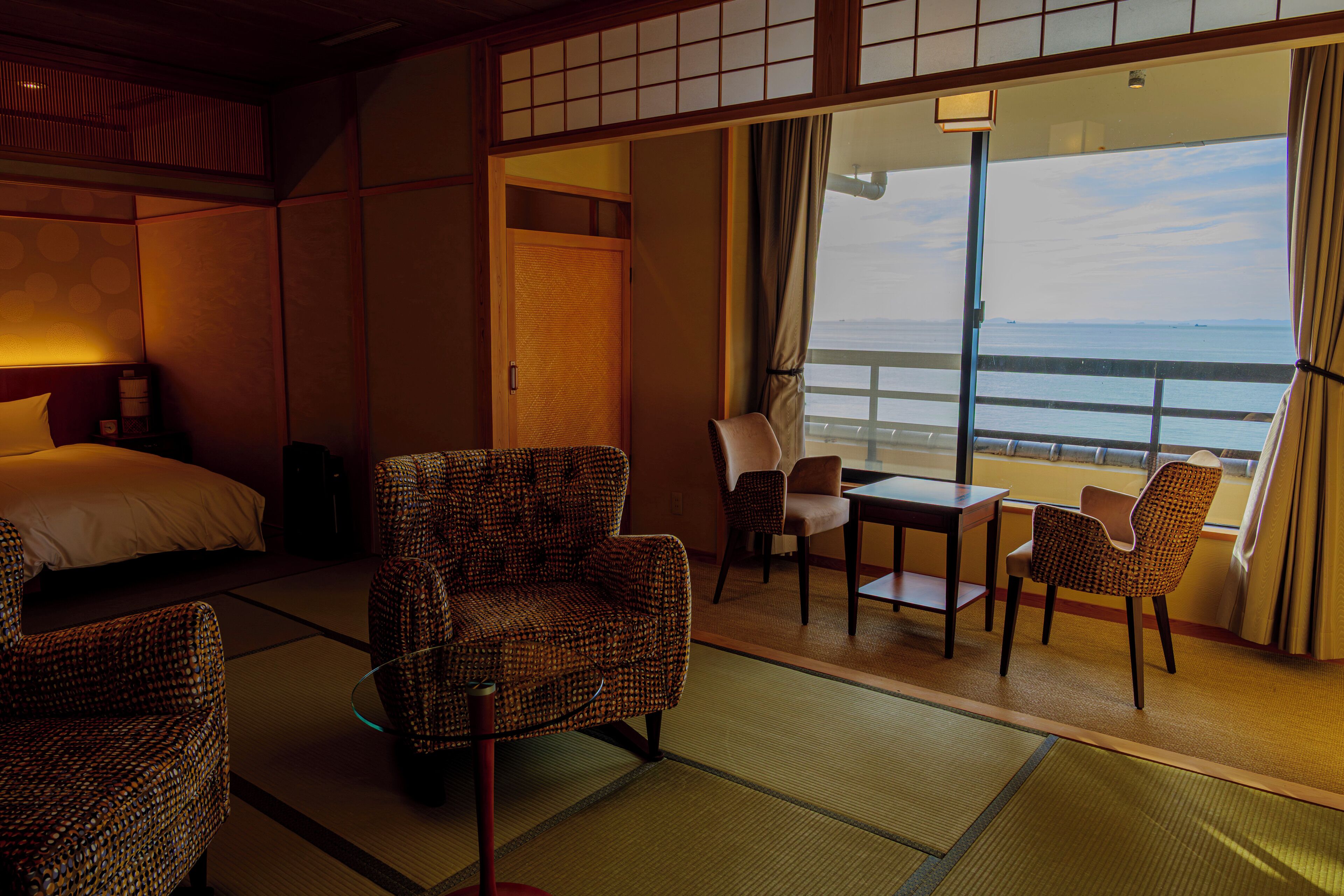 A hotel room with tatami mats, a bed, a dining area with a table and chairs, and a view of the sea.