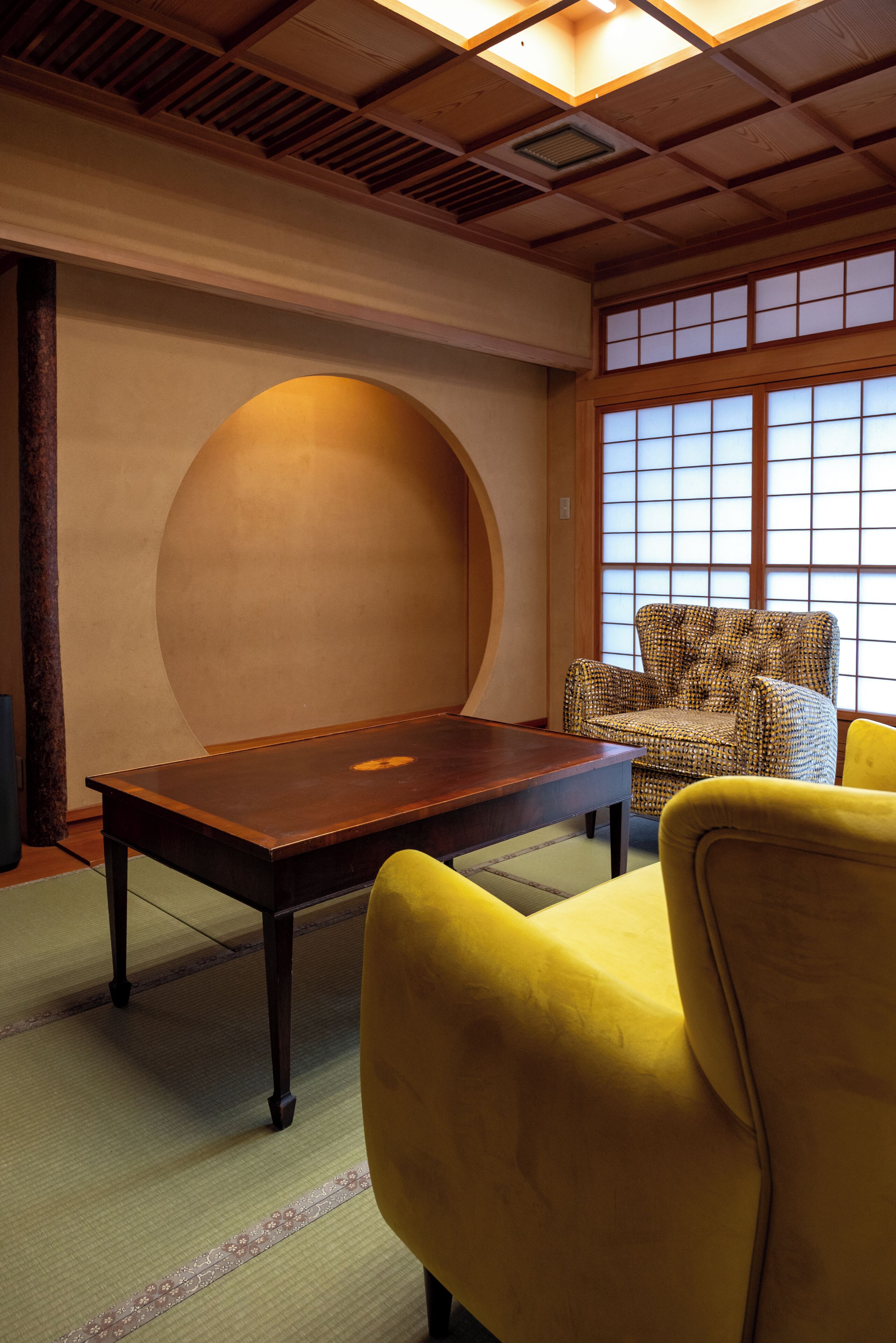 Una habitación japonesa tradicional con suelo de tatami, una mesa de madera baja, un sillón estampado y una ventana con puertas shoji.