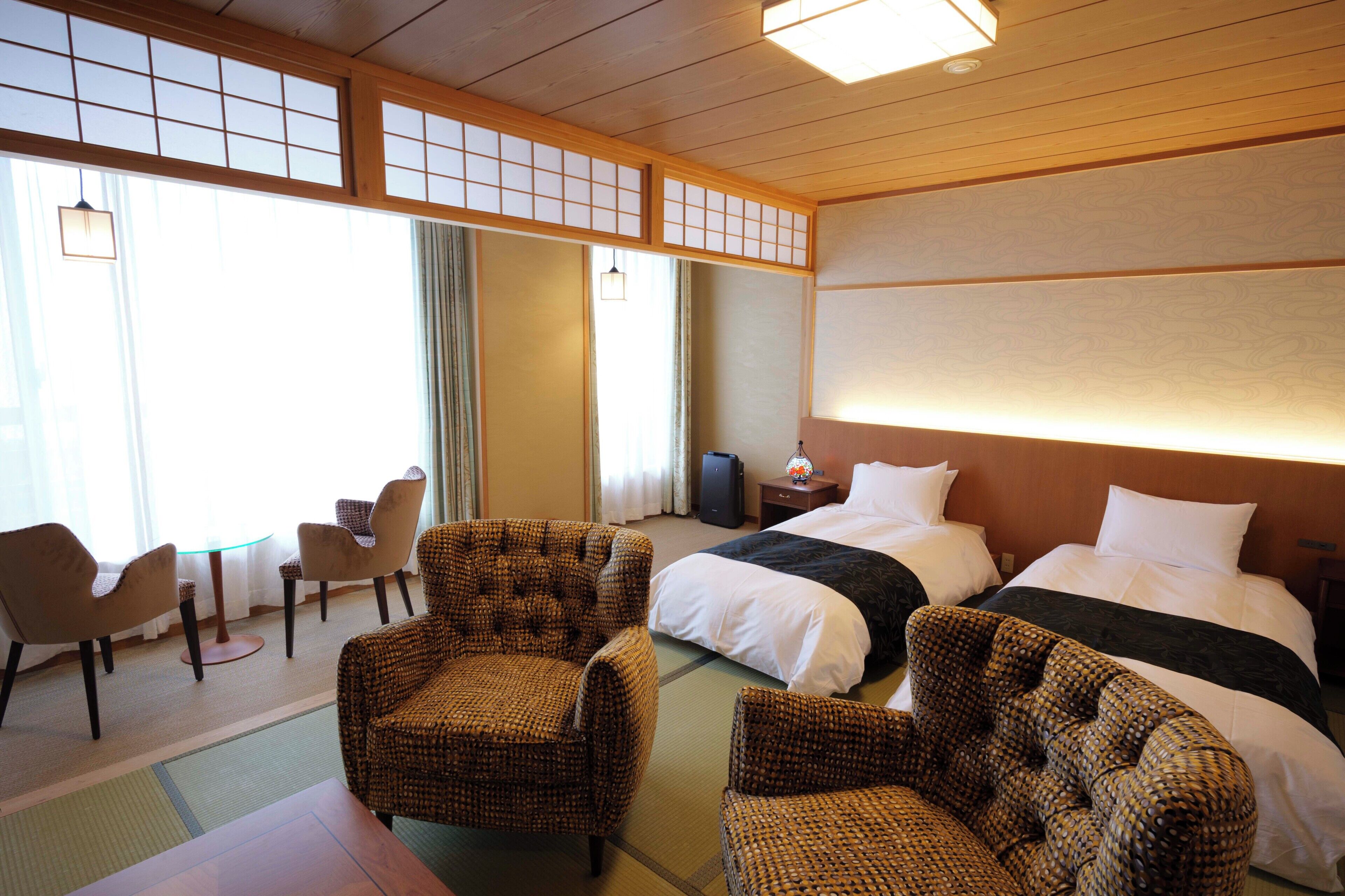 A hotel room with two beds, a sitting area with chairs, a small table, and a view of the outside through large windows.