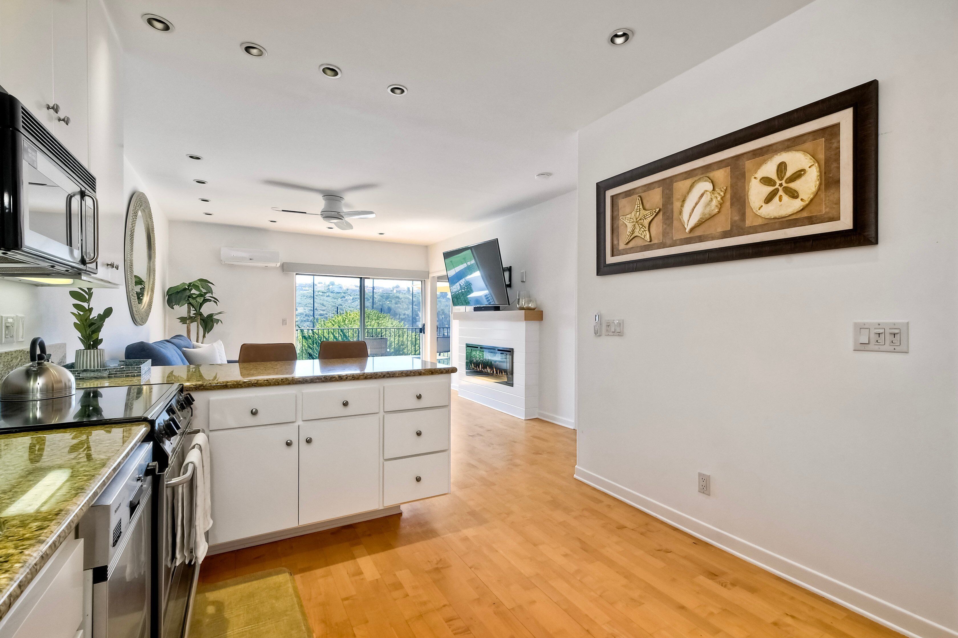 A modern kitchen with white cabinets, a granite countertop, and a built-in oven. There is a dining area with a wooden table and chairs, and a large window offering a view of the outdoors.