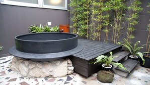 Un cuenco de piedra circular con una plataforma de madera, rodeado de plantas en macetas y un suelo de baldosas.