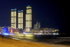 Un paisaje urbano nocturno con rascacielos iluminados y una playa en primer plano.