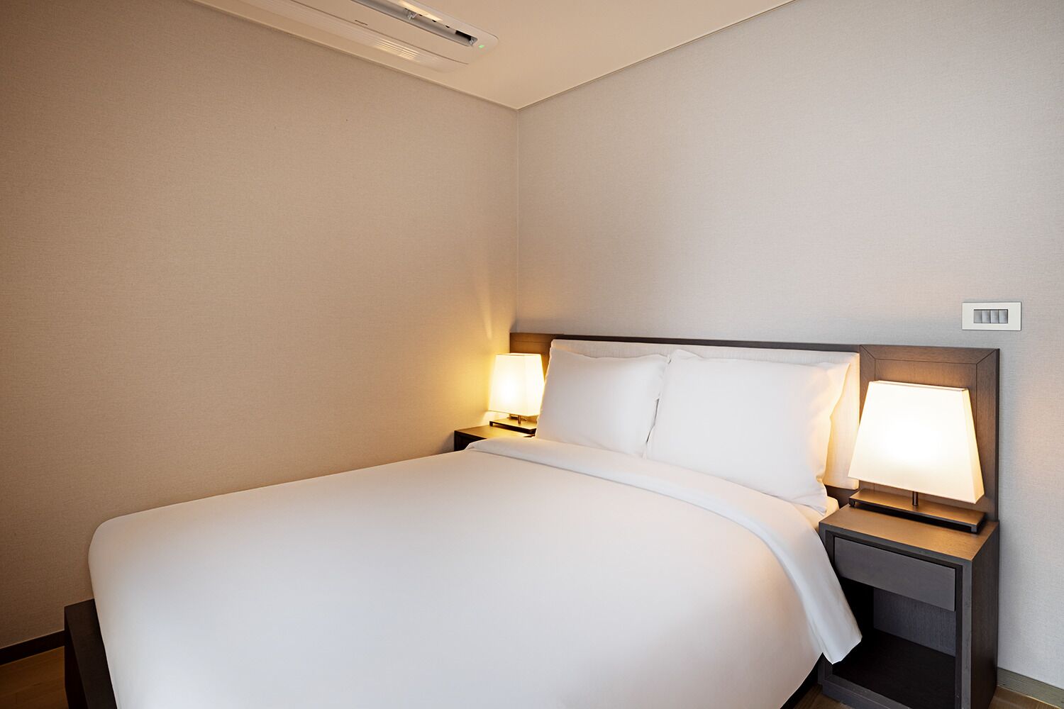A neatly made bed with white linens, two bedside tables with lamps, and a light switch on the wall.