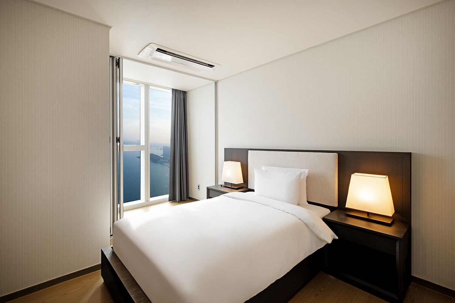 A hotel room with a large bed, two bedside tables with lamps, a window with curtains, and an air conditioning unit.