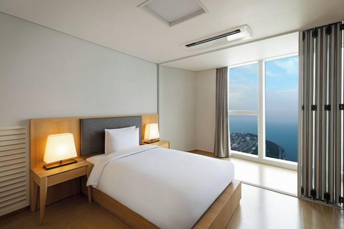 Une chambre d’hôtel moderne dotée d’un grand lit, de deux lampes de chevet, d’une vue sur la ville grâce à de grandes fenêtres du sol au plafond et d’une porte coulissante.