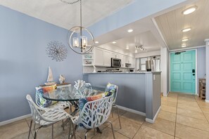 Une cuisine moderne avec des meubles blancs, une porte de couleur turquoise et une table à manger en verre, accompagnée de chaises blanches.