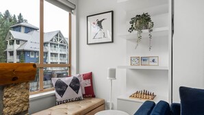 Una habitación acogedora con vistas a una casa a través de una ventana, un tablero de ajedrez, un banco y un sofá azul.