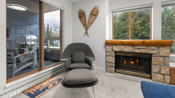 Una sala moderna con chimenea de piedra, un sillón gris confortable y un amplio ventanal con vistas a una zona boscosa.