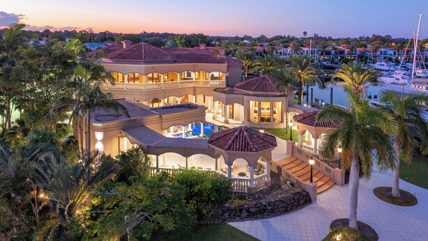 Una casa grande de varios pisos con piscina, rodeada de palmeras y un puerto deportivo.