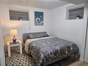 Een slaapkamer met een bed, een nachtkastje met een lamp, een kamerbreed tapijt met een patroon en twee wandplanken met planten.