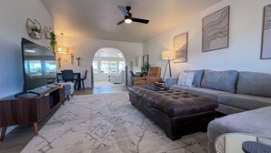Un salon spacieux avec un grand téléviseur à écran plat, un pouf en cuir marron, un canapé gris et un tapis à motifs.