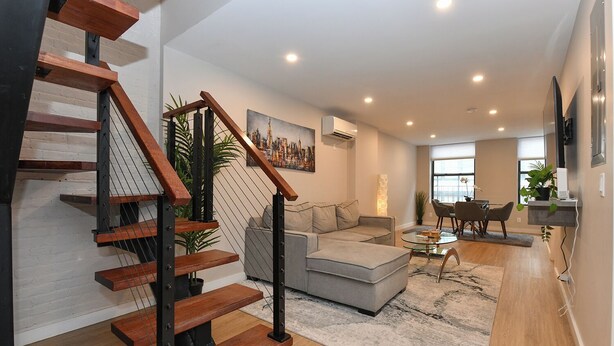A modern living room with a staircase, a grey sofa, a coffee table, and a dining area with chairs. - Elegant SoHo 5BR Duplex – Upscale, Central & Spacious (New York)