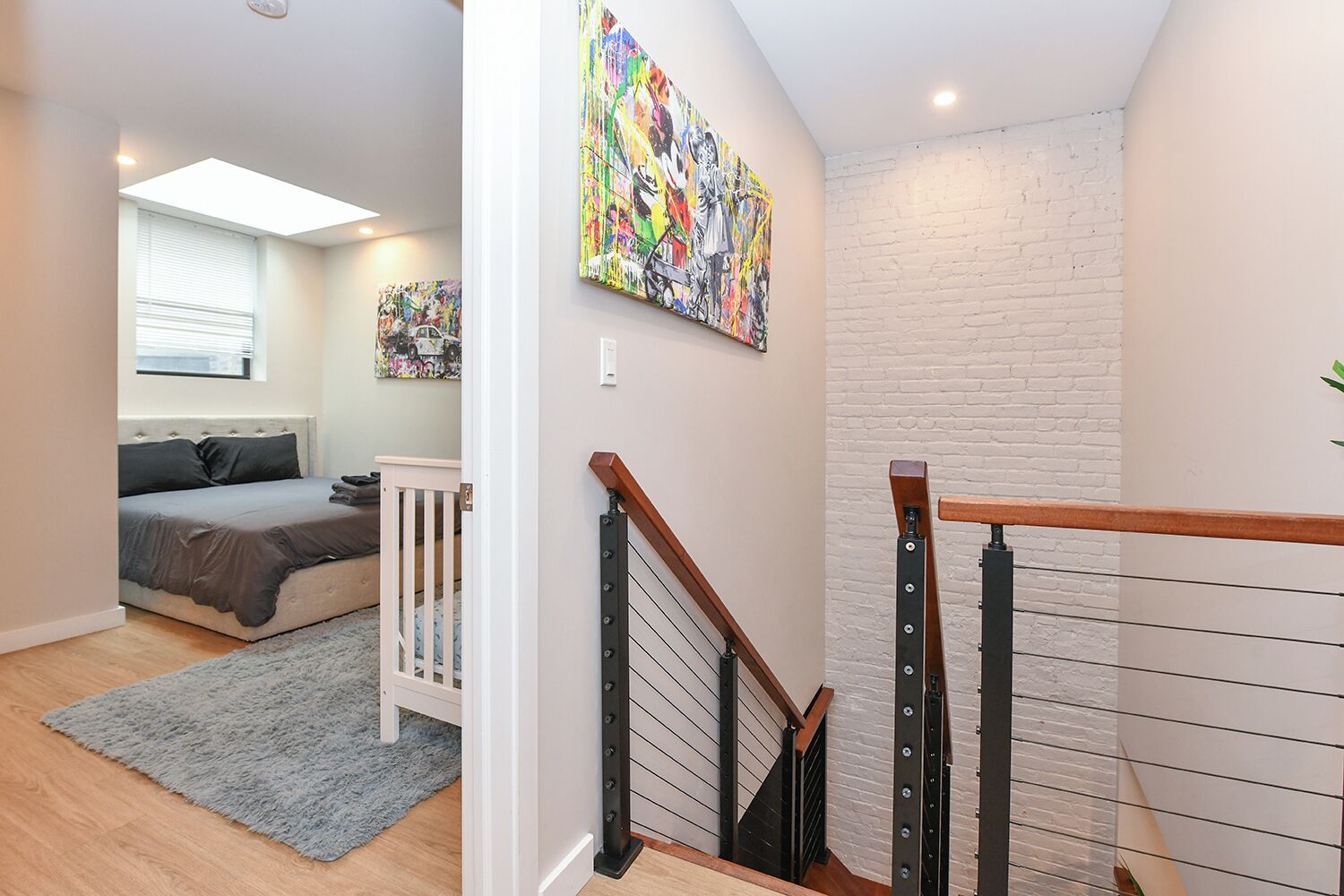 A bedroom with a bed, a gray rug, a white crib, and a staircase with a wooden handrail.