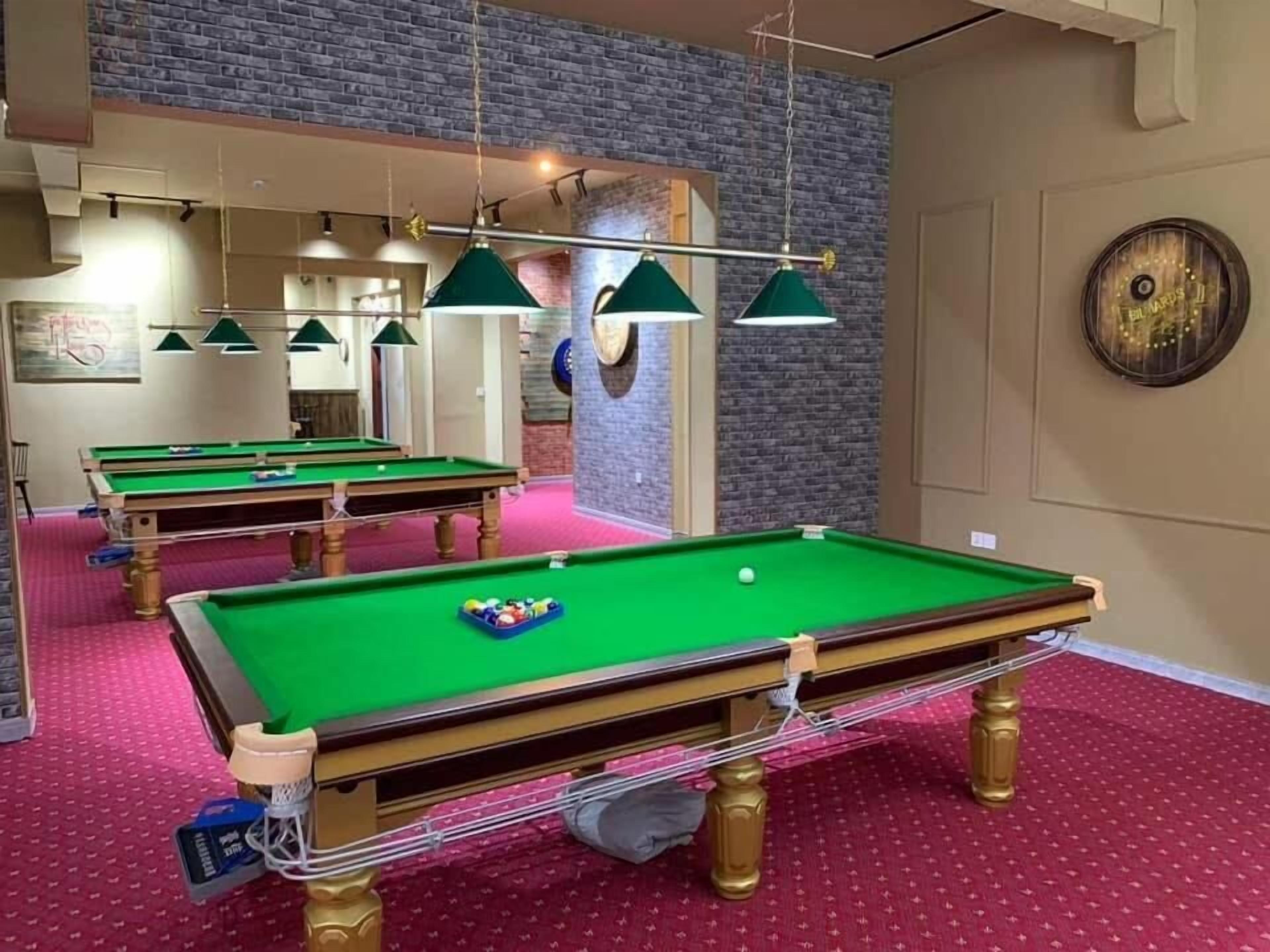 Une salle de billard dotée de plusieurs tables de billard, de tables recouvertes de feutre vert et de lampes vertes suspendues au plafond.